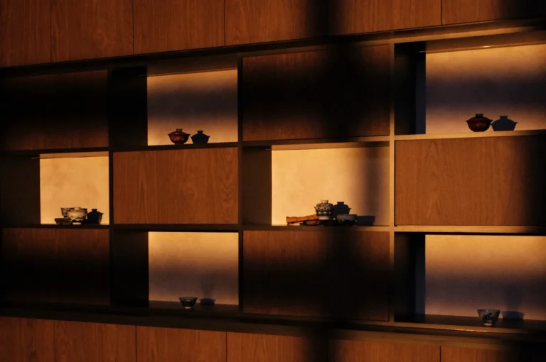 Wooden shelves with dim lighting showcase various small teapots and bowls, casting soft shadows to create a serene, minimalist ambiance.