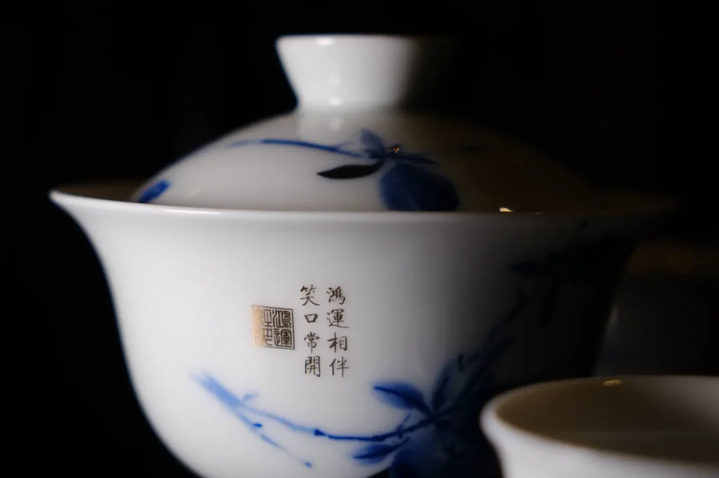 A white ceramic tea cup with a lid, adorned with blue floral designs and Chinese characters, against a dark background, exuding elegance and tranquility.