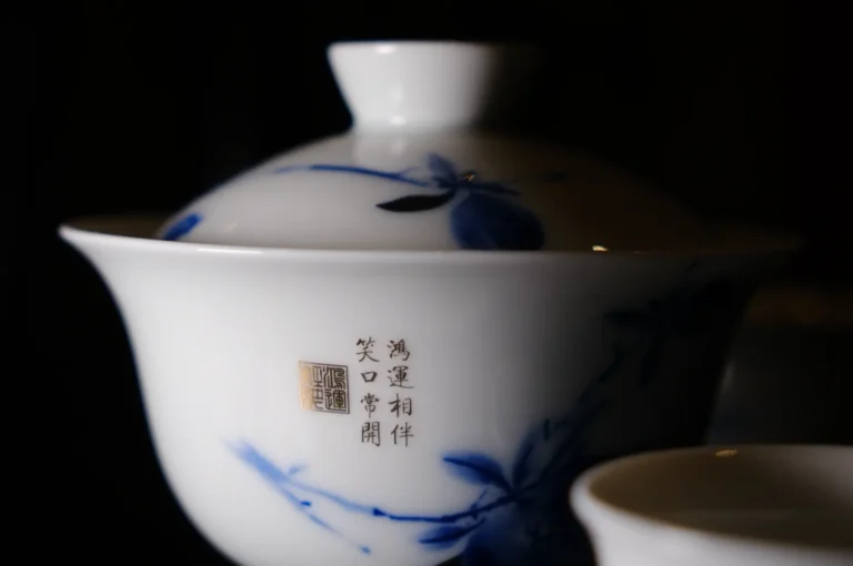A white ceramic tea cup with a lid, adorned with blue floral designs and Chinese characters, against a dark background, exuding elegance and tranquility.