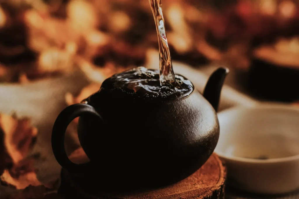 A teapot on a wooden coaster is filled with steaming liquid, surrounded by soft-focus autumn leaves. The scene conveys warmth and coziness.