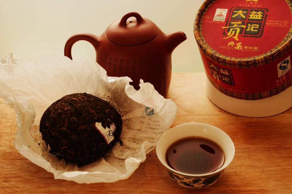A round block of Puerh tea on crumpled paper, a brown teapot, and a cup filled with dark tea sit on a wooden surface, evoking a sense of tranquility.