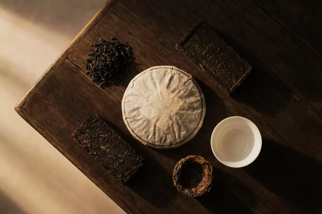 Assorted teas on a wooden table include loose leaves, compressed cakes, and bricks, with a white porcelain teacup, all in warm, natural light.