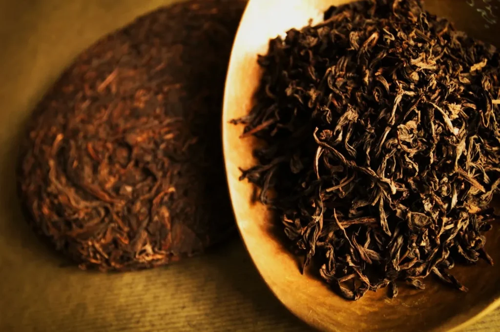 A wooden bowl holds loose leaf tea, rich in dark browns, beside a compact cake of similar texture. The warm tones evoke a calming, earthy ambiance.