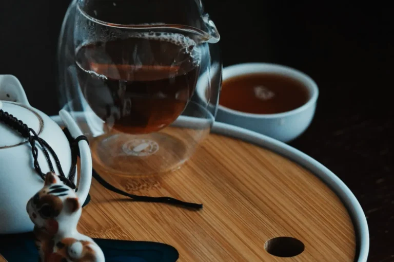 A clear glass teapot with tea sits on a wooden tray. A ceramic cat charm and a small cup in the background create a cozy, serene atmosphere.