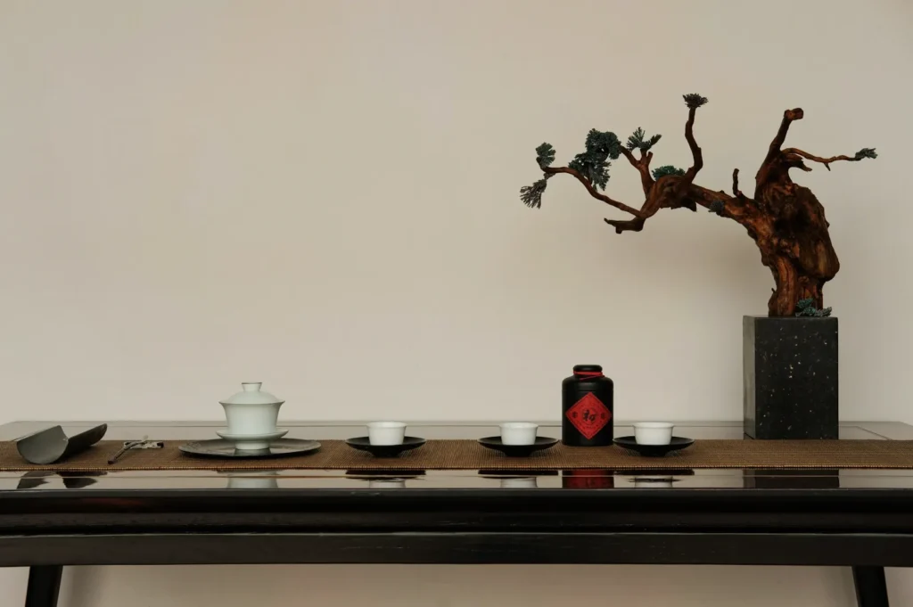 Minimalist tea setup on a dark wooden table with white cups, a black tea canister, and a bonsai tree in a gray pot, conveying tranquility.