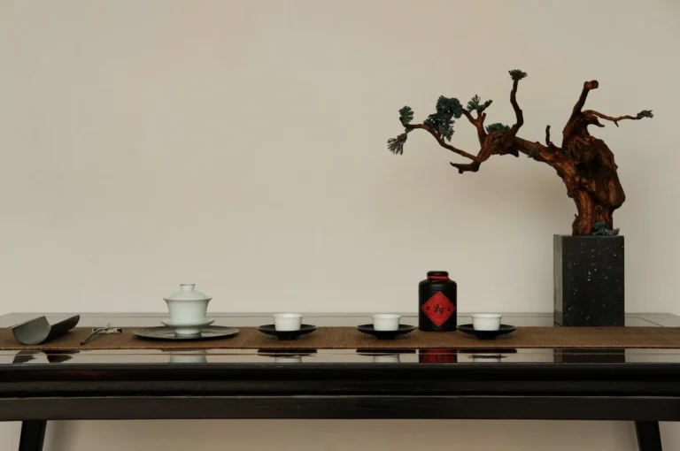 Minimalist tea setup on a dark wooden table with white cups, a black tea canister, and a bonsai tree in a gray pot, conveying tranquility.