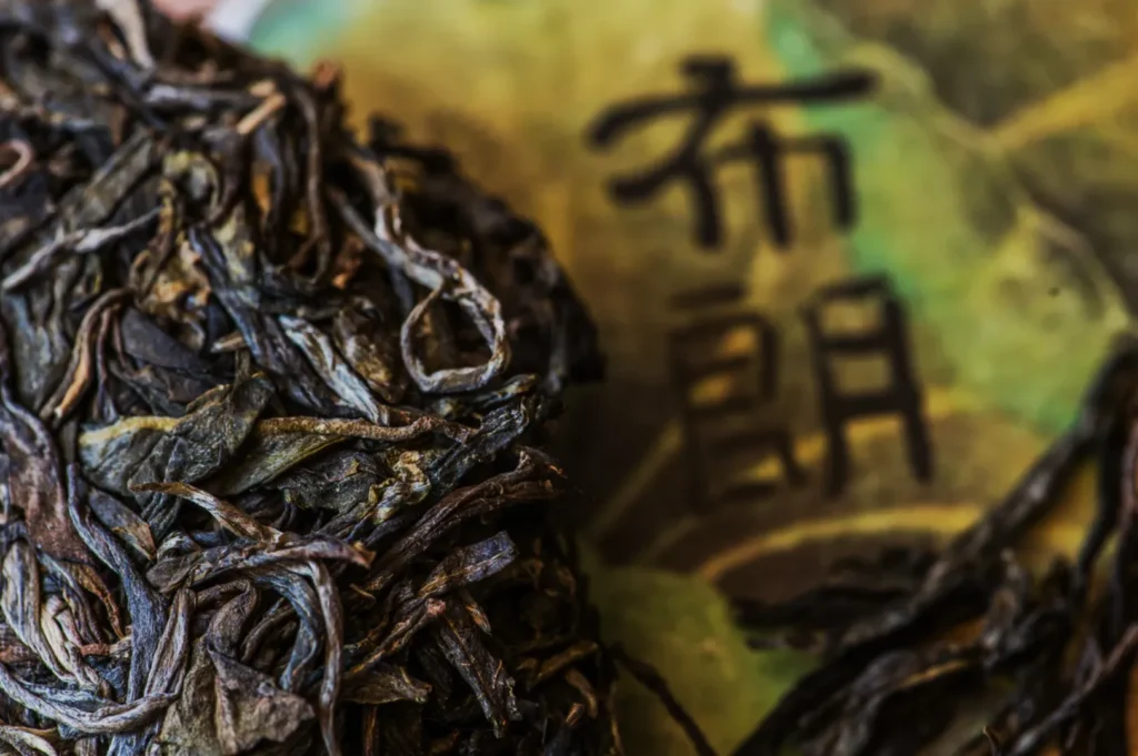 Close-up of dried tea leaves against a blurred background featuring Asian characters. The texture of the leaves appears rustic and earthy.