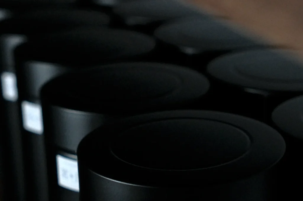 Dark cylindrical objects are lined up in rows, casting subtle shadows. The image has a moody, minimalistic feel with a focus on smooth surfaces.
