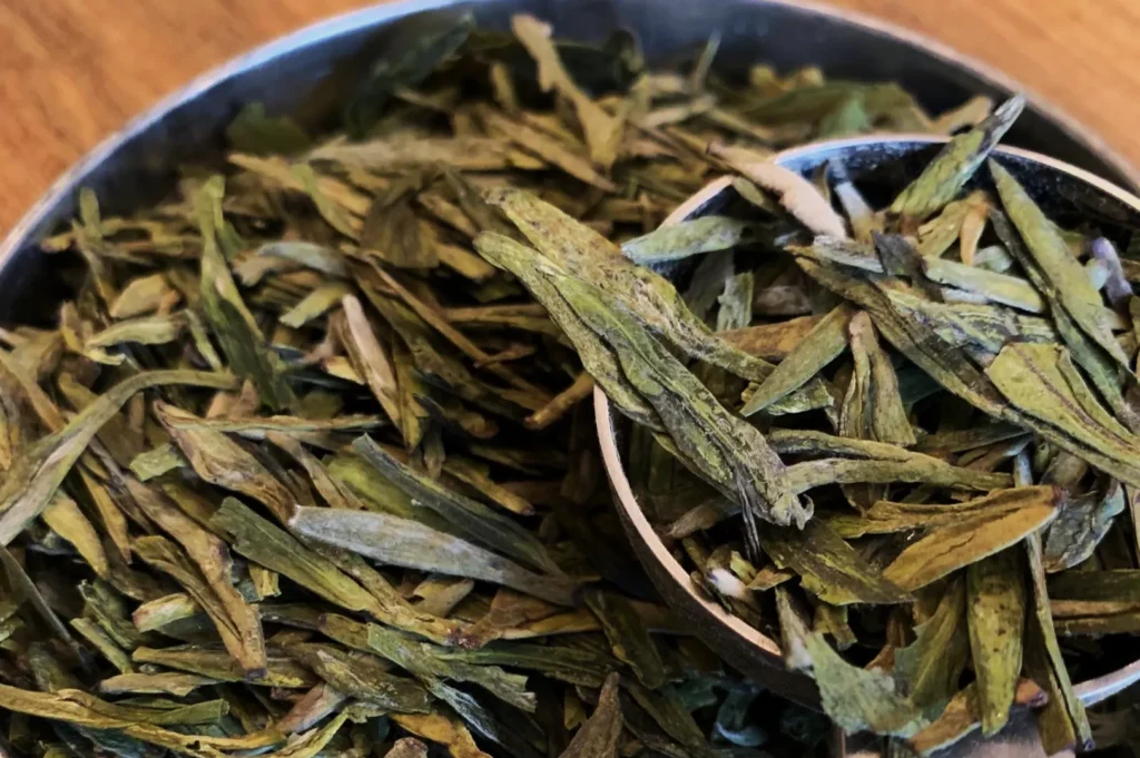 Close-up of dried green tea leaves in a metal container, showcasing their elongated shape and earthy green tones, conveying a sense of freshness.