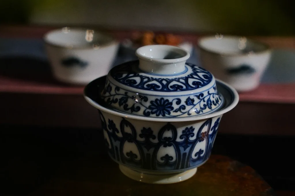 Ornate blue and white porcelain tea bowl with lid, featuring intricate floral patterns. Two blurred cups in the background, creating a tranquil, elegant ambiance.