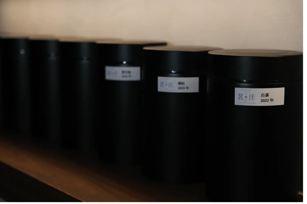 A row of black cylindrical containers with Chinese labels sits on a wooden shelf. The containers' minimalist design creates a calm, organized atmosphere.