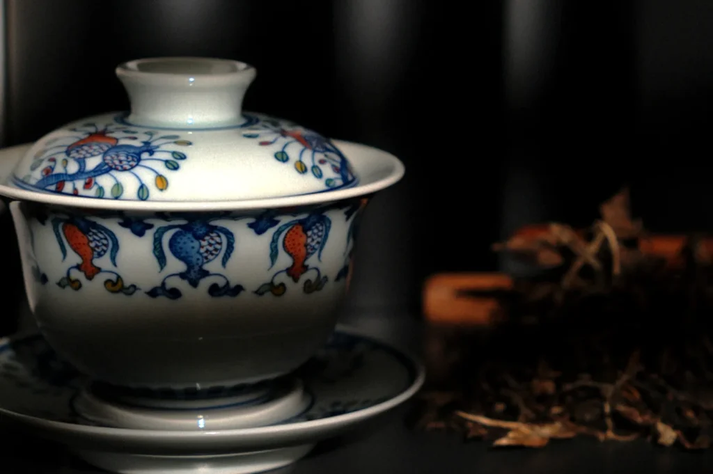 A decorative white ceramic tea cup with lid and saucer, featuring colorful floral patterns sits in soft lighting. Dried tea leaves are blurred in the background.