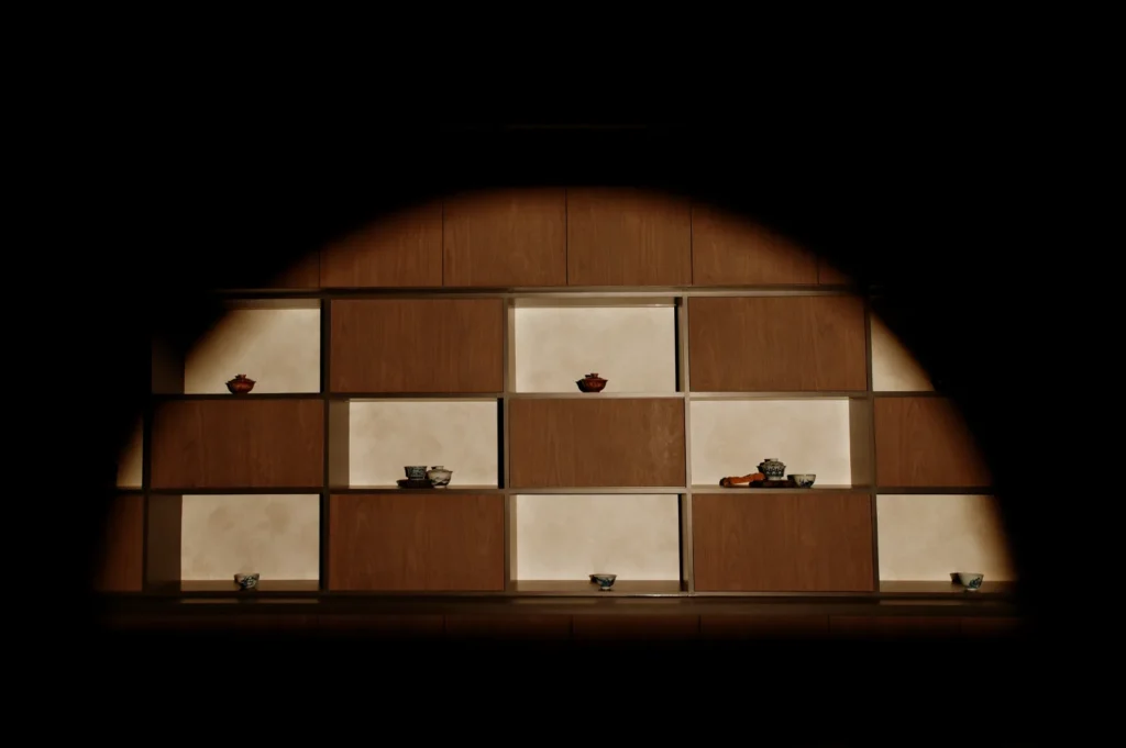 Dimly lit shelves with a geometric design display small ceramic pieces and bowls. The warm, minimalist setting conveys a serene, contemplative mood.