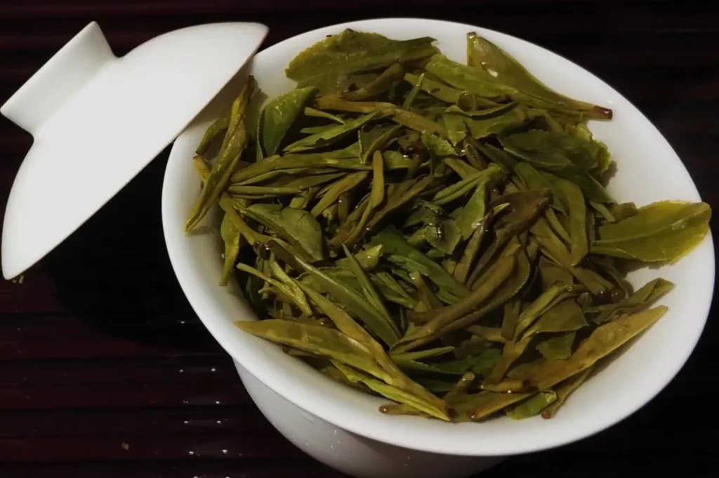 A white ceramic bowl with a lid holds freshly steeped green tea leaves. The leaves are vibrant and glossy, resting on a dark wooden surface.