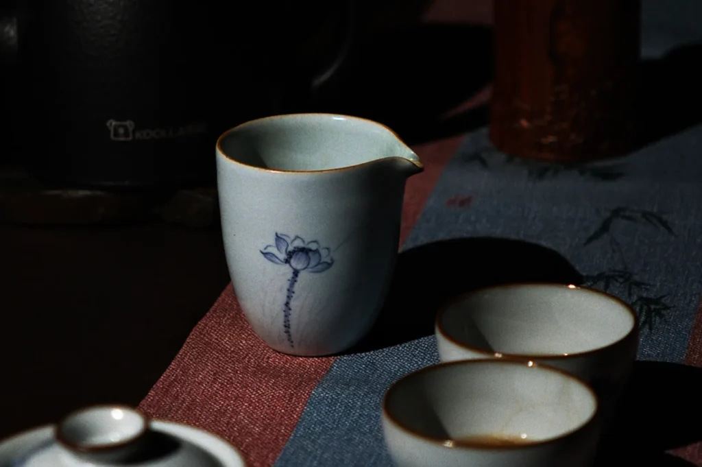 A tranquil scene features a small white ceramic pitcher with a blue lotus design, set on a blue and red tablecloth, surrounded by tea cups. Soft lighting creates a calm atmosphere.