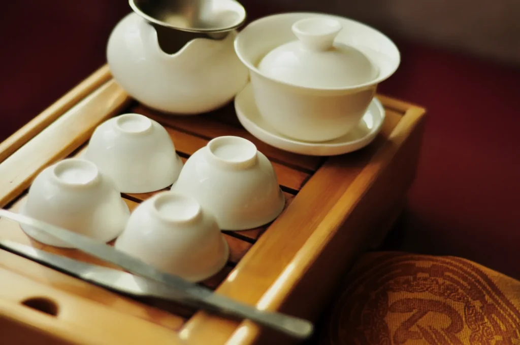 A set of white teacups, a teapot, and saucer rest elegantly on a wooden tray. A cloth with a decorative pattern lies nearby, evoking a serene tea ritual.