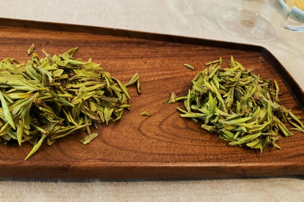 Two small piles of fresh green tea leaves sit on a rectangular wooden tray, placed on a light-colored tablecloth. The setting is calm and inviting.