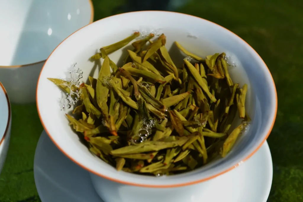 Close-up of a white ceramic bowl filled with green tea leaves steeping in hot water. Small bubbles are visible, creating a fresh, calming ambiance.