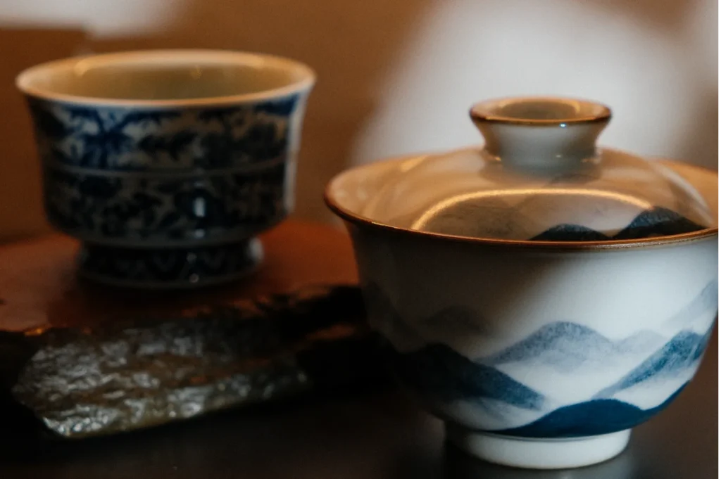 Close-up of two elegant ceramic cups on a stone surface. A blue and white floral cup and a lidded bowl with mountain motifs create a serene, artistic atmosphere.