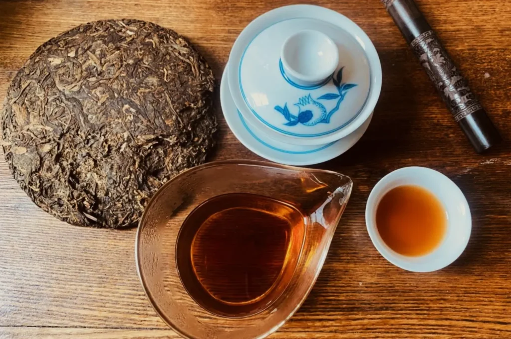 A rustic wooden table holds a round Pu-erh tea cake, a clear glass pitcher with amber tea, a small white cup, a blue-patterned gaiwan, and a dark cylindrical tea holder. The scene is calm and inviting.