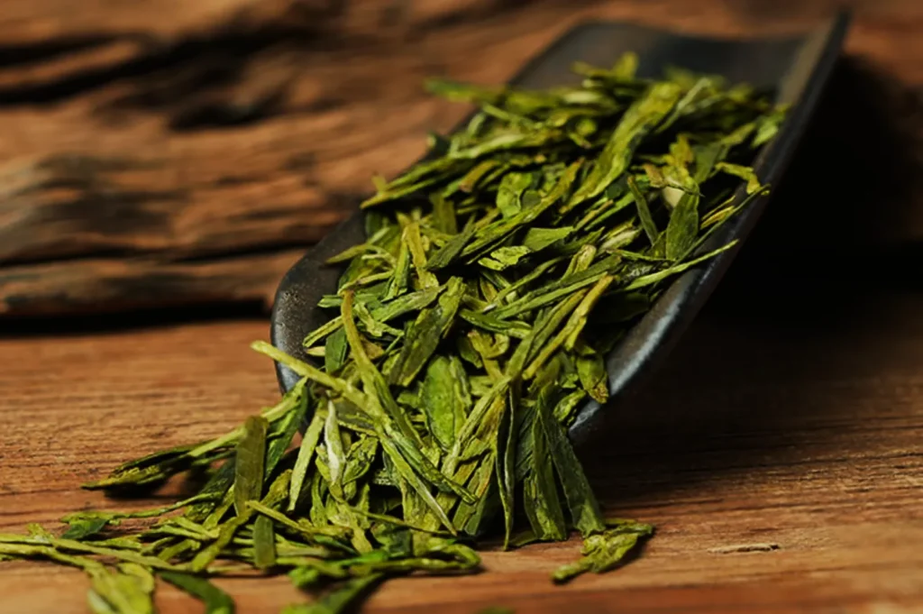 Loose green tea leaves on a dark wooden scoop rest on a textured wooden surface, evoking freshness and natural simplicity.