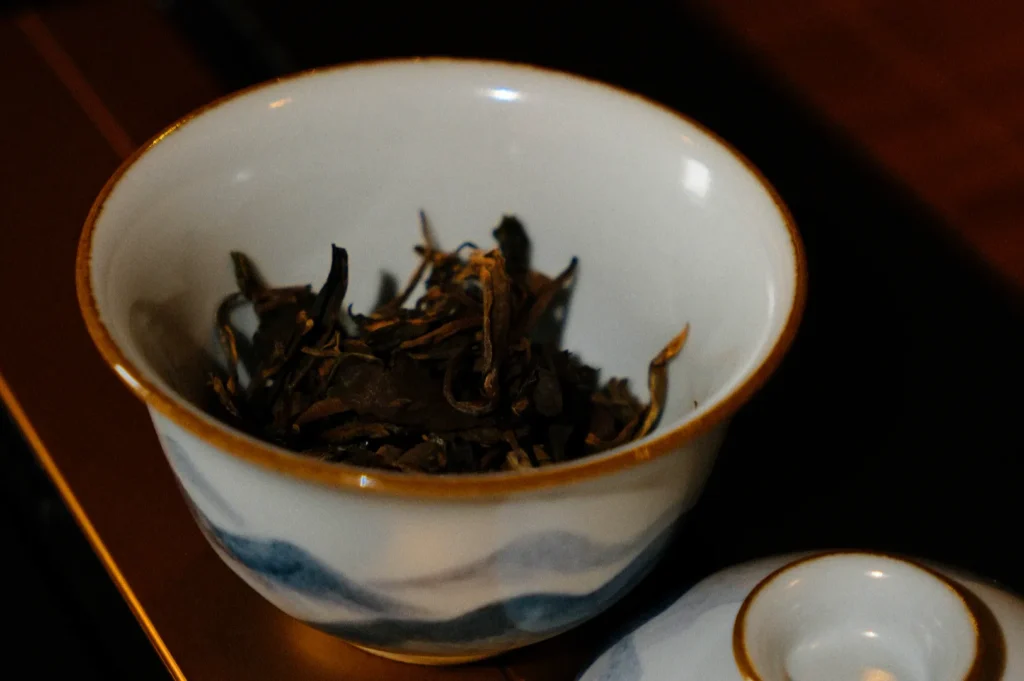 A decorative bowl with a gold rim and blue pattern, filled with loose leaf tea. A matching lid rests beside it on a wooden surface, giving a cozy feel.