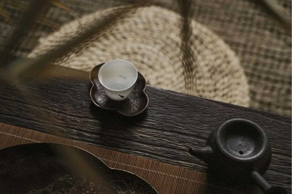 Minimalist tea setting on a wooden table with a small black teapot, a white cup on a flower-shaped saucer, and a woven mat in the background. Calm ambiance.
