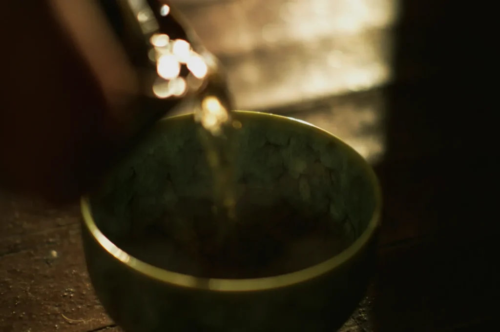 Warm tea being poured into a textured, green ceramic bowl. The lighting is soft and moody, creating a serene and calming atmosphere.