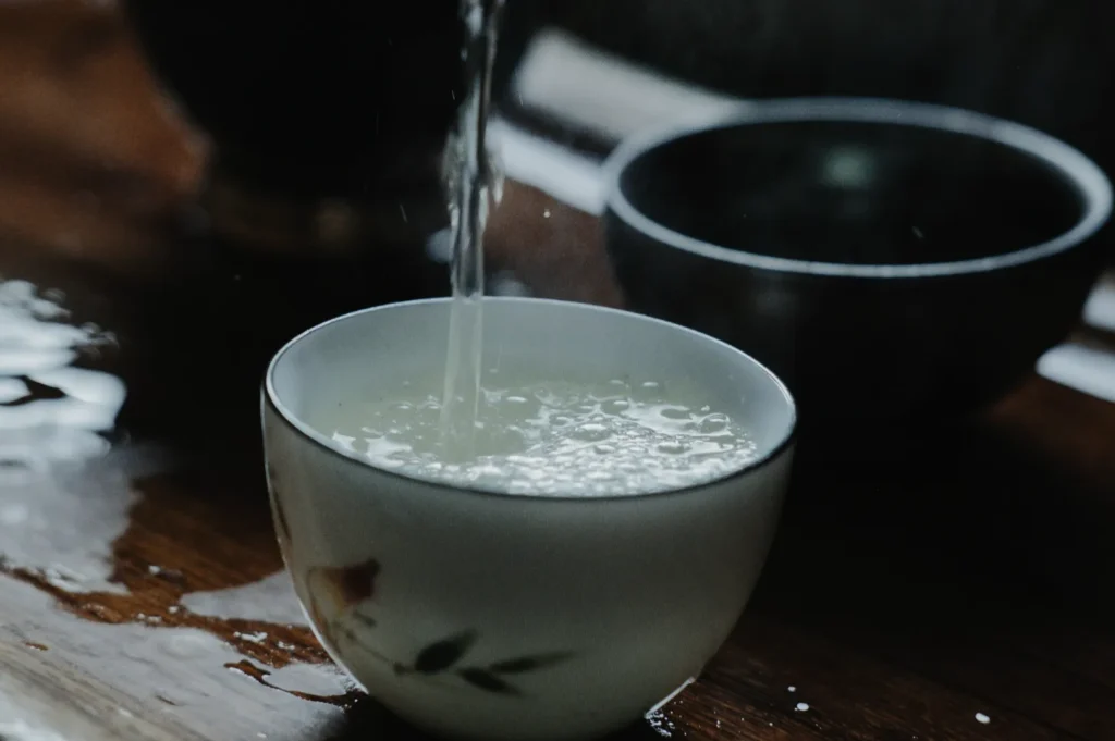 Pouring liquid into a white ceramic cup with a floral design, creating ripples. A dark bowl is beside it on a wooden table. The mood is calm.