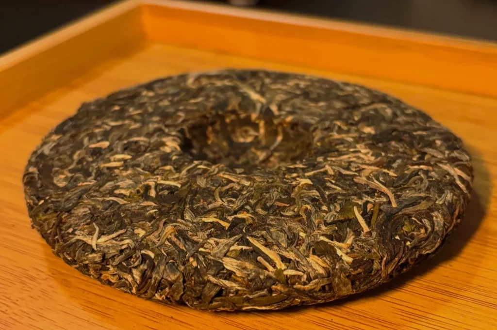 A round, compressed pu-erh tea cake with a dark, rich texture sits on a wooden tray, suggesting an earthy aroma and traditional tea experience.