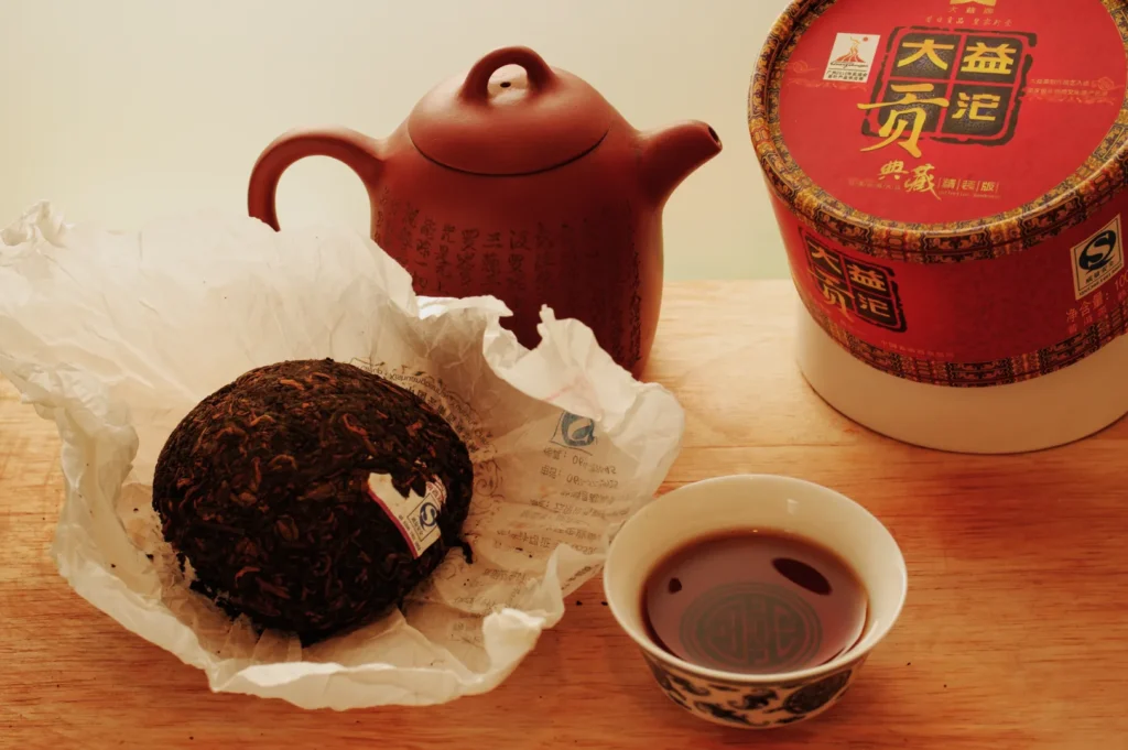 A pressed tea brick on paper, a brown clay teapot, and a Chinese tea cup with dark tea are on a wooden surface. A round tea box is nearby.