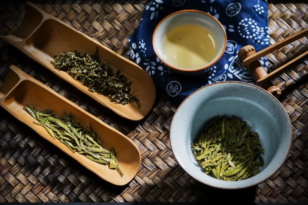Assorted loose leaf teas on bamboo trays, a ceramic bowl, and a cup of brewed tea on a woven mat. The scene conveys a calm and traditional ambiance.