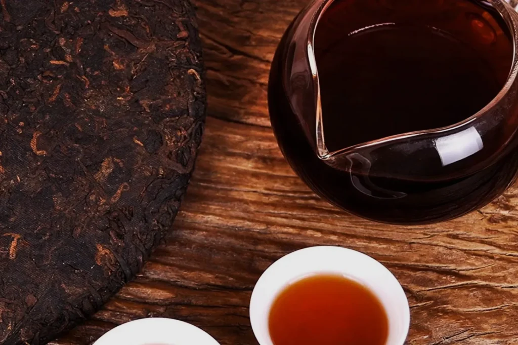 A rustic wooden table displays a round block of compressed tea, a clear glass teapot with dark tea, and two small white cups filled with brewed tea.