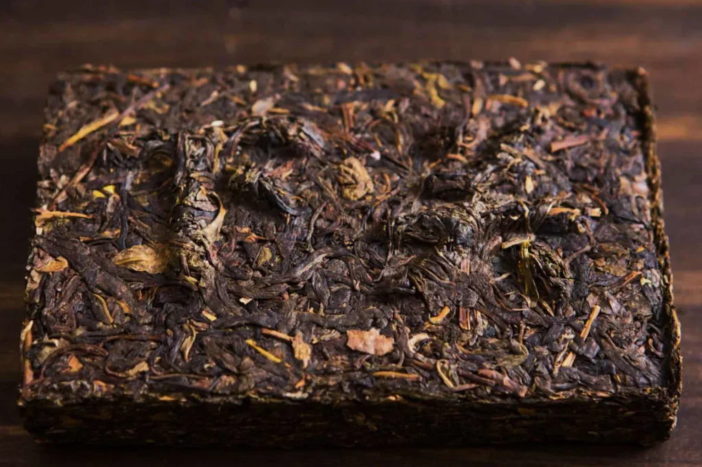 A dense, rectangular block of compressed, dark-brown tea leaves sits on a wooden surface. The texture is rough, with visible twigs and leaves.