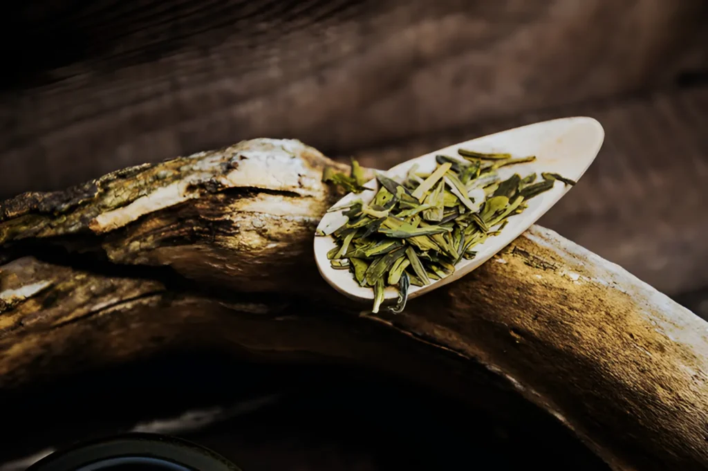 A spoonful of dried green tea leaves rests on a rustic wooden surface. The earthy tones convey a natural and calming atmosphere.