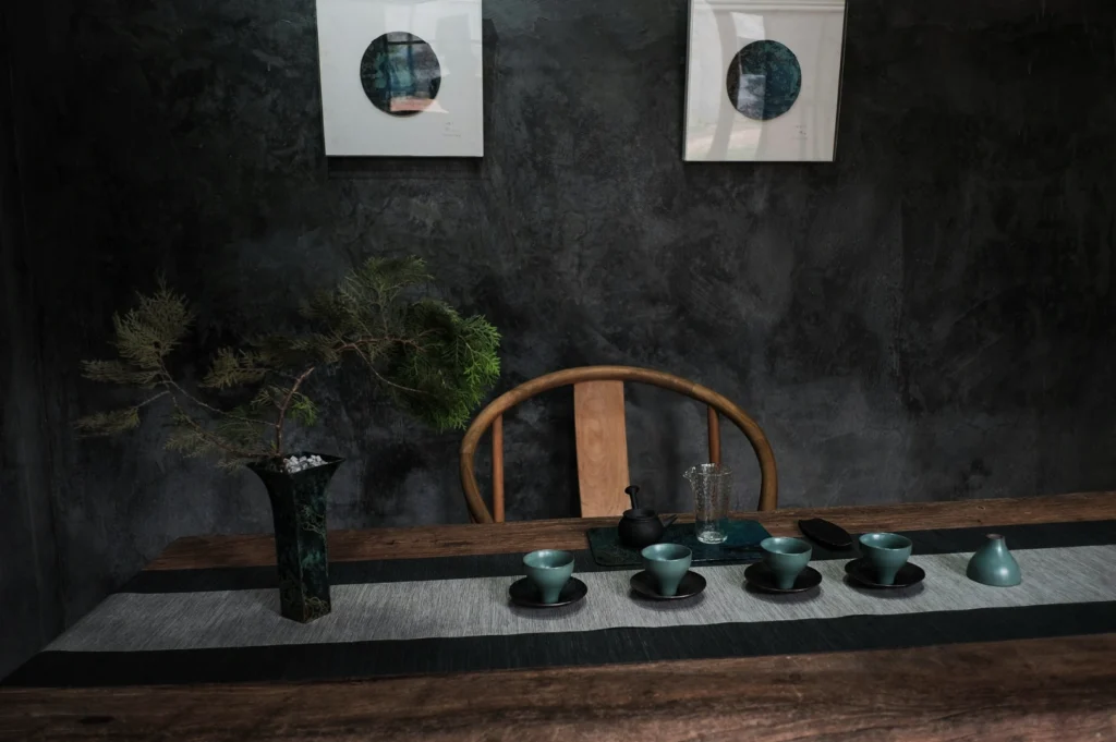 Minimalist tea setup on a wooden table with green cups, teapot, and vase holding a branch. Two abstract artworks hang on a gray wall. Serene atmosphere.