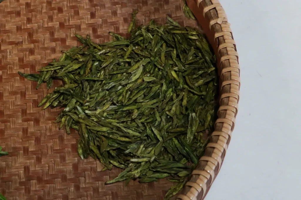 A woven basket partially filled with dried green tea leaves on a white surface. The texture of the basket and leaves creates a natural, organic feel.