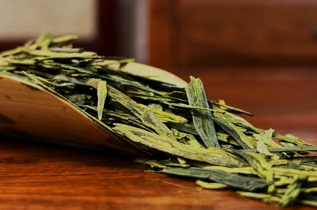 Loose green tea leaves spilling over from a wooden scoop onto a smooth, warm-toned wooden surface, evoking a sense of natural freshness and calm.
