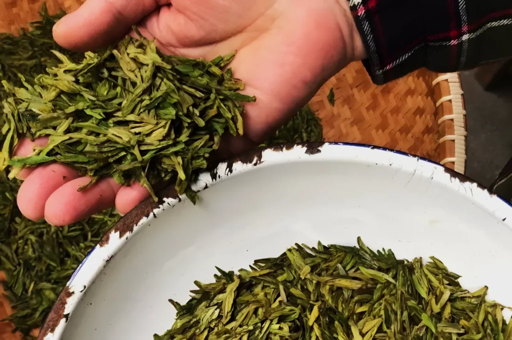 A hand holds a bunch of green tea leaves over a white bowl filled with more leaves. A woven basket is partially visible, creating an earthy, natural tone.