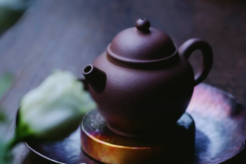A small, brown teapot sits on a metallic tray with a soft focus white flower in the foreground. The scene conveys a calm, serene atmosphere.