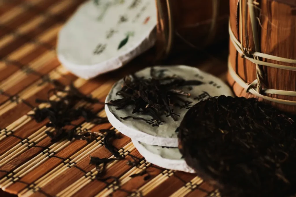 Pressed tea cakes and loose tea leaves on a woven bamboo mat. Wooden containers beside them suggest a rustic, traditional setting.