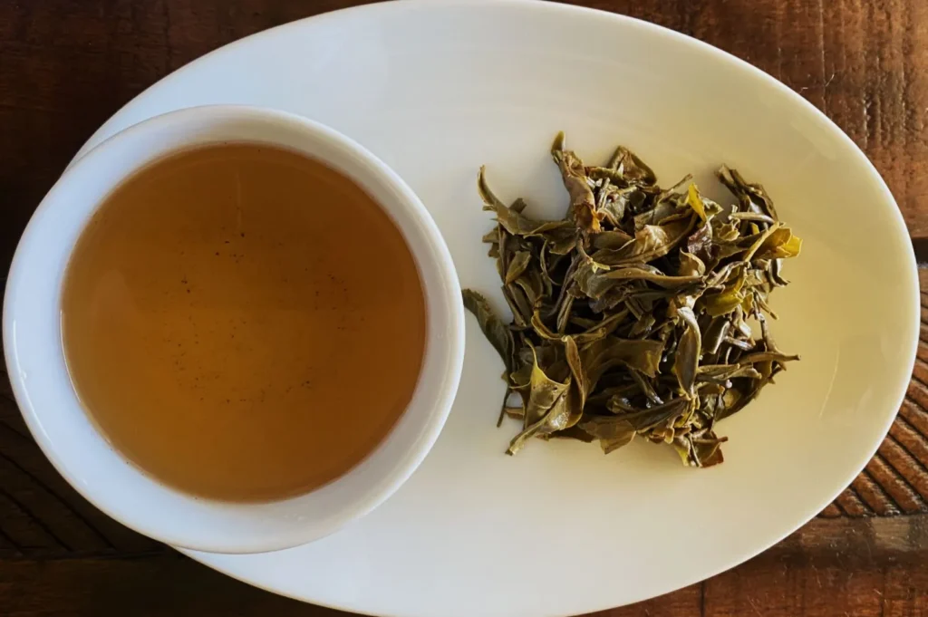 A white cup with light amber tea and a pile of dried tea leaves are on an oval white plate. The setting rests on a wooden surface, creating a warm, rustic vibe.