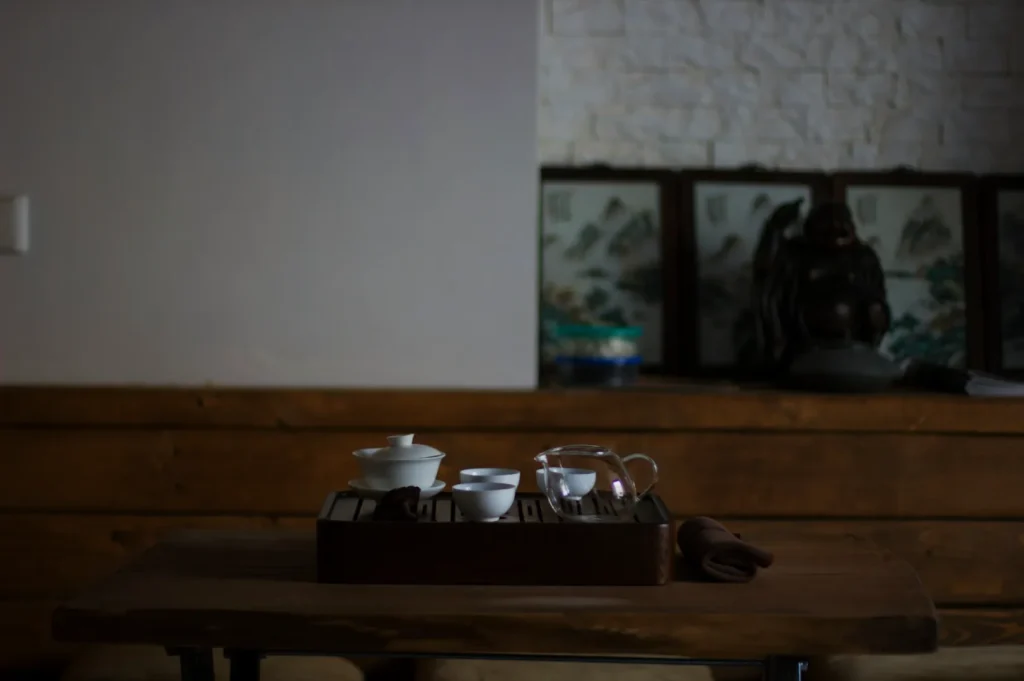 A wooden table with a traditional tea set, including cups and a teapot, is in focus. The background features decorative panels and a Buddha statue, exuding a serene atmosphere.