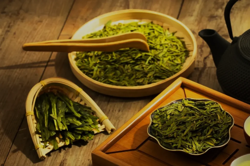 A display of vibrant green tea leaves in various containers, including a bamboo basket and round tin on a wooden table, next to a black teapot. Cozy ambiance.