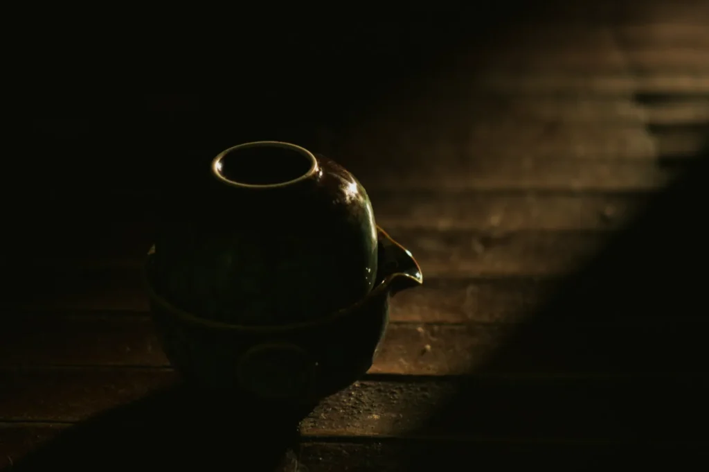 A dimly lit ceramic teapot sits on a wooden table, casting a soft shadow in the warm, focused light, creating a tranquil and serene atmosphere.