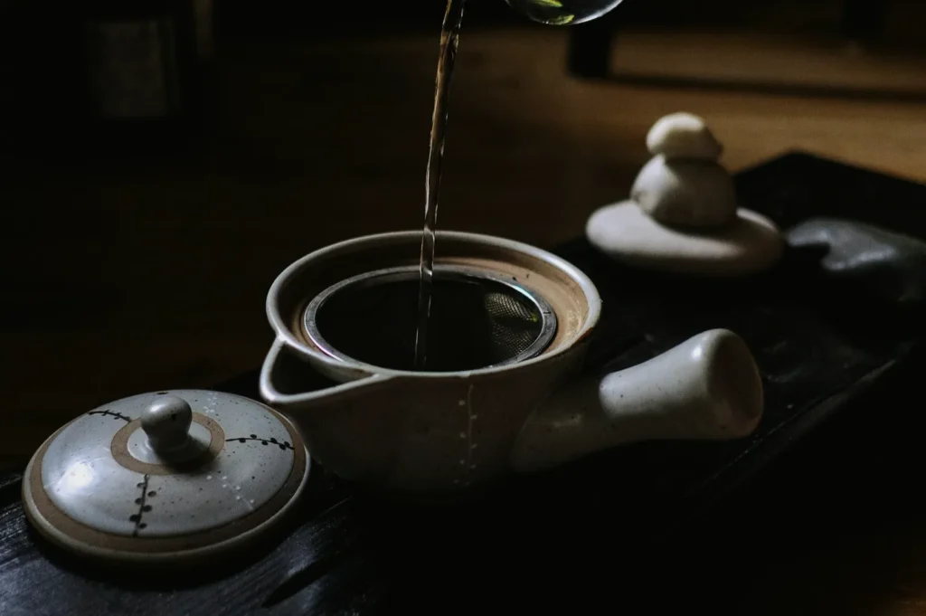 A close-up of water being poured into a ceramic teapot with a metal infuser. The lid and a stack of rounded stones are nearby, creating a serene ambiance.