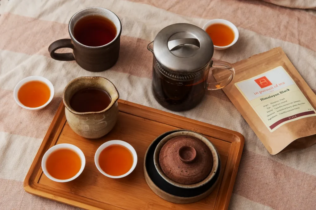 A cozy tea setup on a striped cloth featuring a teapot, cups, and a package labeled "Himalayan Black." Warm tones suggest relaxation.