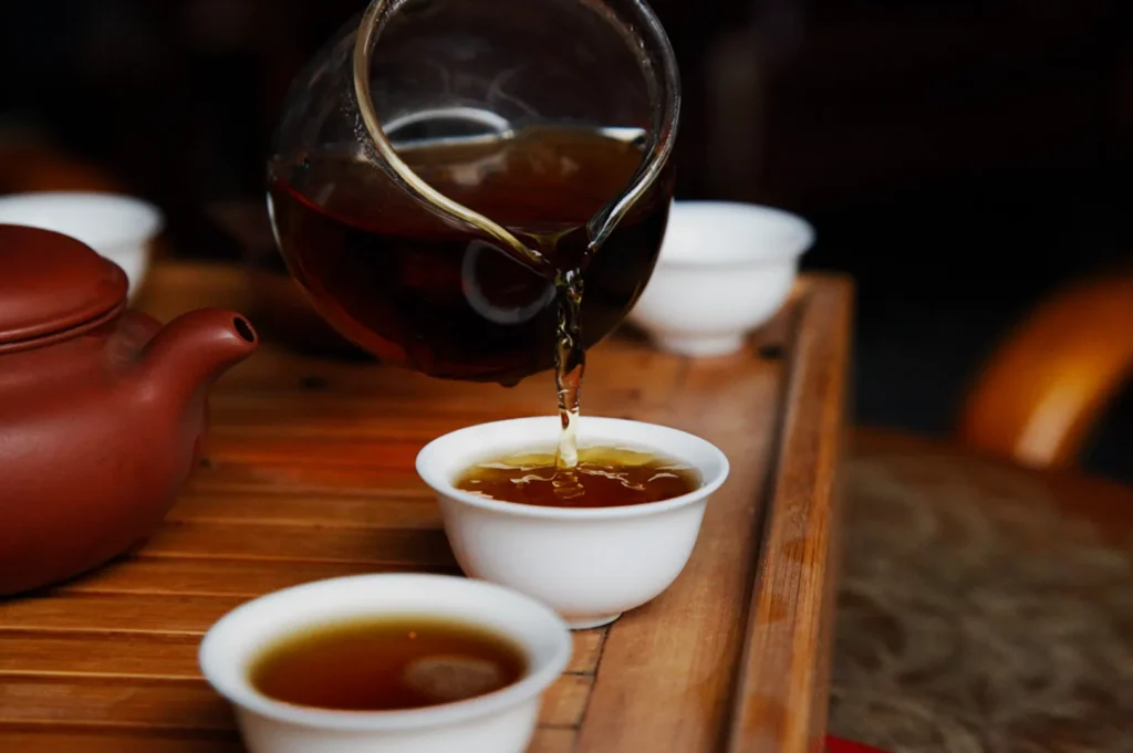 A teapot pours amber tea into a small white cup on a wooden tray, creating a warm and tranquil atmosphere.