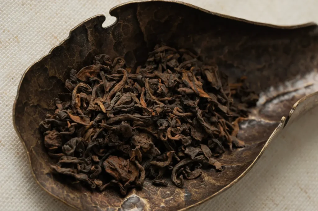A pile of dried tea leaves rests in a textured, metallic scoop on a light fabric background. The scene conveys a rustic, organic feel.