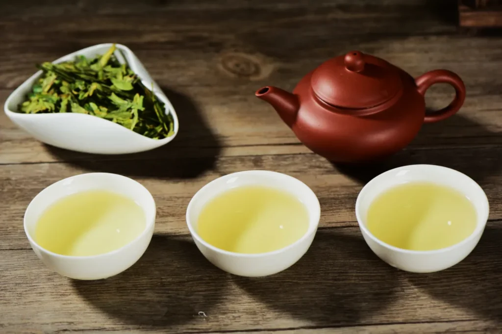 Brown teapot with three cups of light green tea on wooden table; white bowl of fresh green tea leaves beside. Calm, inviting atmosphere.